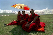 Three monks with umbrella sitting in green open landscape watching festival