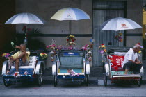 General.  Street scene with three bicycle rickshaws decorated with flowers and flags parked beside roadside kerb with resting drivers.