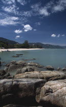 View over rocks and bay of turquoise water towards distant moored boat and quiet sandy beach.