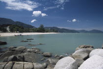 View over rocks and bay of turquoise water towards distant moored boat and quiet sandy beach.