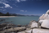 View over rocks and bay of turquoise water towards quiet sandy beach.