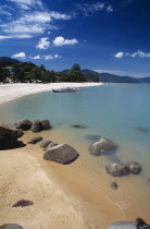 View along shore of quiet sandy beach with moored boat in middle distance and smooth rocks in shallow water in the foreground.