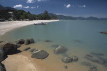 View along shore of quiet sandy beach with moored boat in middle distance and smooth rocks in shallow water in the foreground.