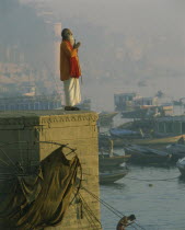 Holy man praying on stone wall above River Ganges with misty town and boats in background