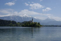 View over the lake toward Bled Island and tower of the Church of the Assumption with snow capped peaks of the Julian Alps behind