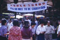 Street scene with mixed crowd below government banner stating Its So Nice To Be Courteous.