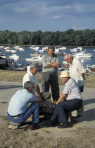 Group of men gathered around playing Chess with boats on The Danube river behind.