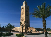 Koutoubia Mosque. Tower seen from pavement with blue sky behind. Palm trees in front.