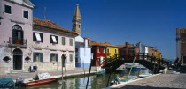 Burano Island.  Bridge and canal with boats moored to posts along each side overlooked by pink  blue  red and yellow buildings and bell tower behind.