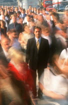 City businessman in suit standing motionless surrounded by crowds in blurred movement.