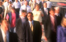 City businessman in suit standing motionless surrounded by crowds in blurred movement.