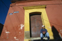 Man in baseball hat sitting on stone step of doorway framed by yellow arch set in red painted wall.