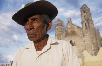 Elderly man in hat  head and shoulders portrait with church building part seen in background.