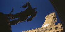 Piazza della Signoria.  Angled view of statue of Perseus with the head of Medusa by Cellini in silhouette with Palazzo Vecchio in sunlight behind.