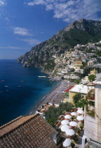 View over balcony with tables and chairs under white sun umbrellas towards beach and village houses built on steep hillside beyond