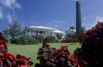 Exterior with people on white painted balcony and old sugar factory tower and wall on the right.  Red and purple variegated plants in the foreground.