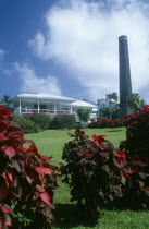 Exterior with people on white painted balcony and old sugar factory tower and wall on the right.  Red and purple variegated plants in the foreground.