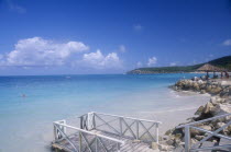 Raised area of sand fortified by rocks with people sitting on loungers under thatched umbrella with man swimming and wooden jetty in the foreground.