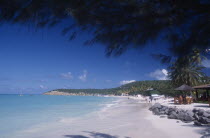 View along beach towards tree covered headland with people walking along shoreline and sitting under thatched sun umbrellas nearby.