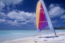 Empty sandy beach with hobiecat with brightly coloured sail in the foreground.  Flat calm aquamarine sea and cloudscape behind.