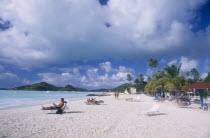 View along sandy beach lined with palm trees and sun umbrellas towards hilly coastline.  People sunbathing on loungers in the foreground.