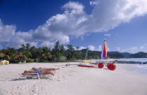 Sandy beach fringed with palm trees with sailing boat and beach-bicycle on the shoreline and three sunbathers on white loungers in the foreground.