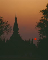 Pha That Luang Sacred Stupa silhouetted against red setting sun and orange sky.