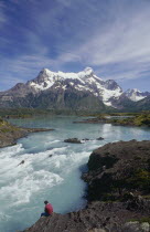 Man sitting on the riverbank beside rapids below the distant snow covered mountains