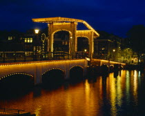 Magere Brug or Skinny Bridge illuminated at night with lights reflected in the canal below.