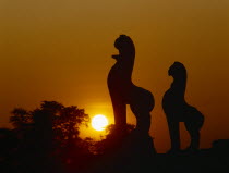 Angkor Wat.  Golden setting sun beyond silhouetted trees and statues.