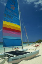 Catamaran with sail up on beach with tourists sunbathing and walking by water