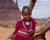 Portrait of Navejo girl wearing a red dress and turquoise necklace and broach leaning against a dead tree trunk.