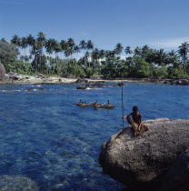 Children in outrigger canoe near beach with boy on rock