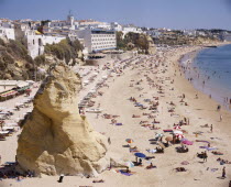 View along the beach from above with a large rock and sunbathers. Hotels and town beyond