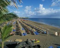 Puerto del Carmen.  Sandy beach with lines of blue sun loungers and orange and green parasols for hire.  Few people sunbathing and hotels beyond.