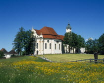 Visitors on road to white painted church with red tile roof and domed clock tower near Steingaden with field of buttercups in the foreground.Bavaria Bayern