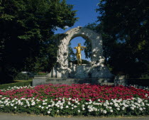 Golden statue of Johann Strauss framed by stone arch in Stadt Park with bed of pink and white geraniums in the foreground.