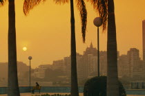 City skyline at sunset with trunks of palm trees in foreground framing single figure on bench.Former portuguese territory reverted to Chinese rule in 1999