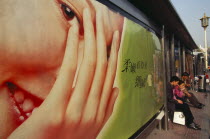 People waiting at bus stop south of Tiananmen Square with advertising hoarding covering exterior wall.Peking Beijing