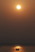 Ganges River at dawn with rowing boat silhouetted against warm sunlight reflected in the water