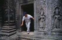 Ta Prohm.  Visitor climbing through doorway of twelth century Buddhist temple with exterior wall decorated with bas relief carvings.Asian Cambodian Kampuchea Religion Southeast Asia History Holidayma...