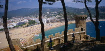 View over busy sandy beach and town from fortified walls of Villa Vella with line of trees in the foreground.  Costa Brava