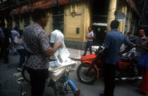 Newspaper vendor selling goods from a bicycle seat in the street with passing traffic and people