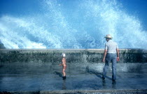 Old man and small boy standing on pavement with waves crashing over the sea wall