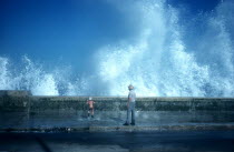 Old man and small boy standing on pavement with waves crashing over the sea wall