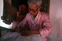 Seated Buddhist Monk writing whilst being fanned by a woman on his right.