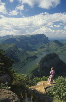 View from 3 Rondavels Point with a woman standing on a flat otcrop of rock looking out at view.