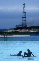 Two men sitting in a shallow swimming pool with Oil derrick in the background