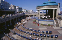 View towards the Promenade Band Stand with a brass band performing on a sunny day and spectators watching from deckchairs layed out in a semi-circle.