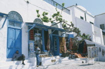 Rugs and postcards for sale.  White painted shopfront with blue doors.  Two local men in shade of awning.